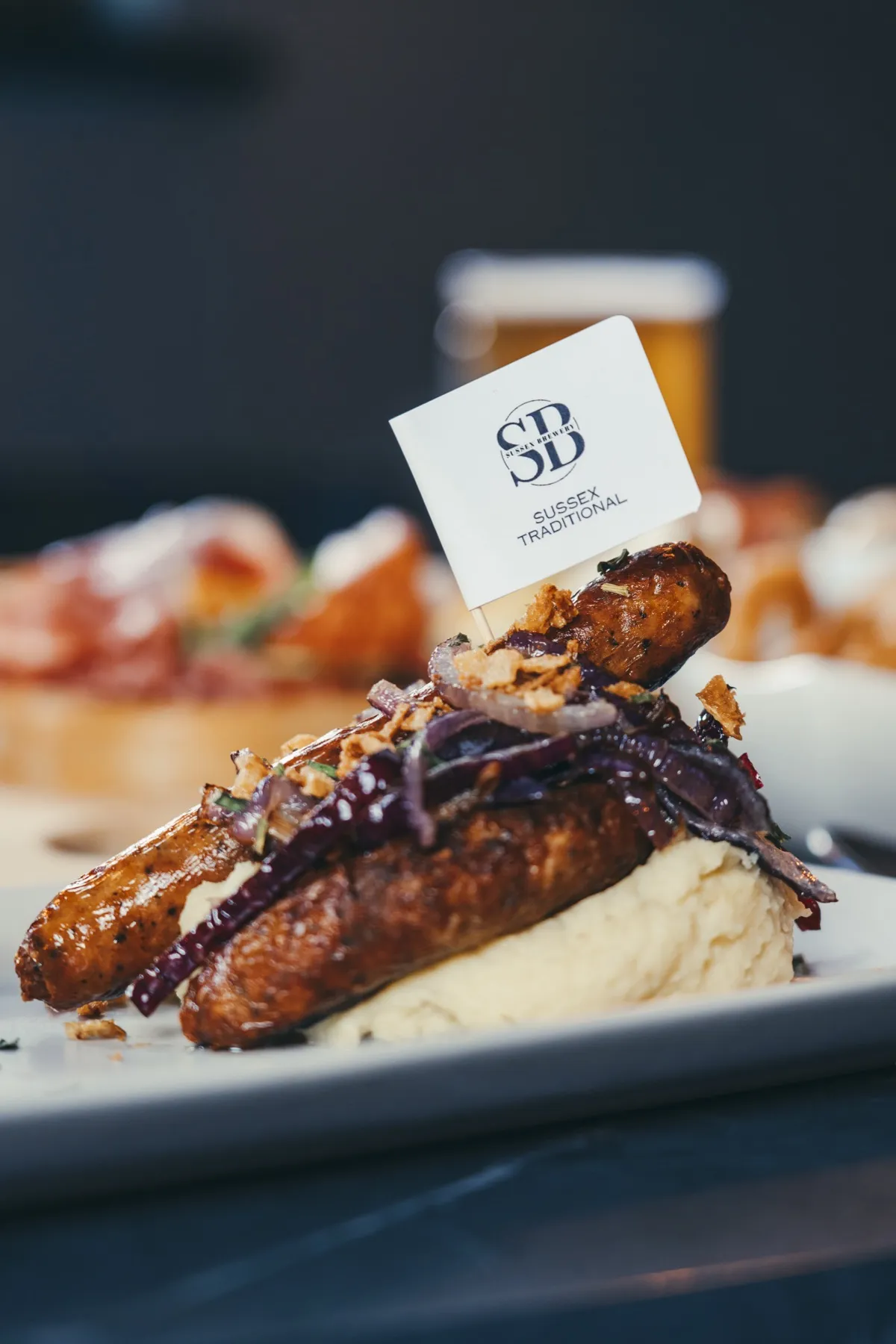 Treagust sausage on mash with caramelised onions and a Sussex Brewery flag